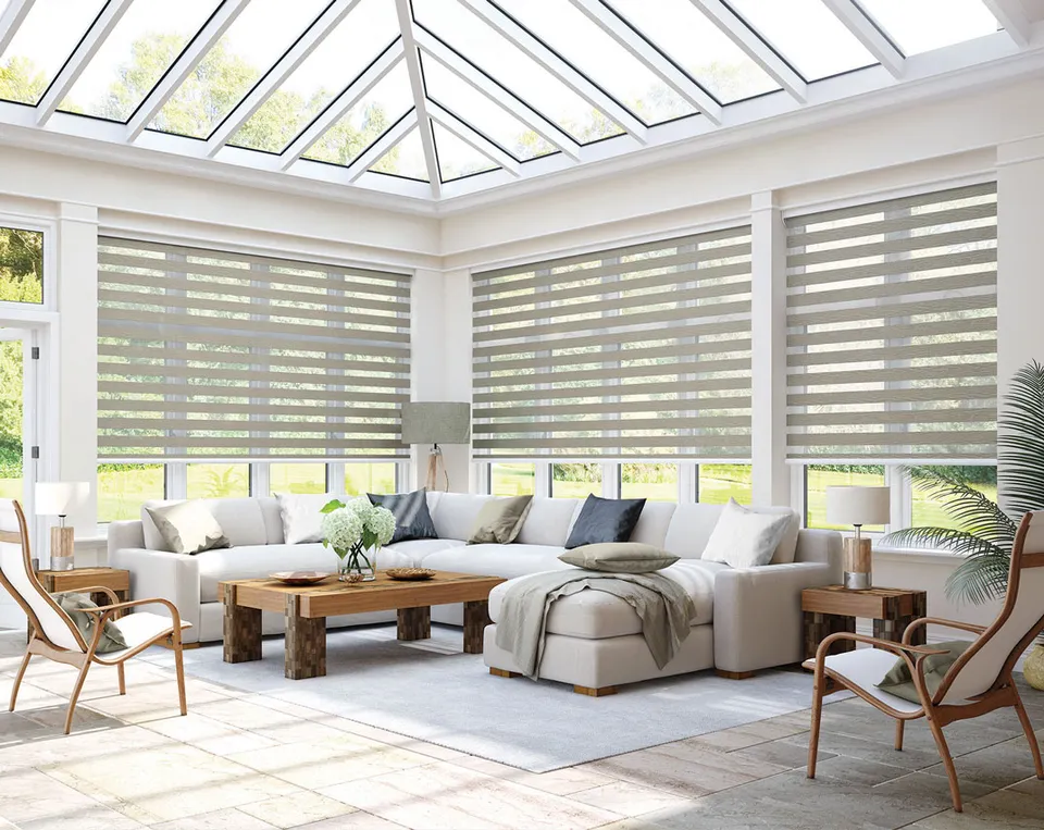 Mink day and night blinds on every window of a bright conservatory with a corner sofa and glass roof