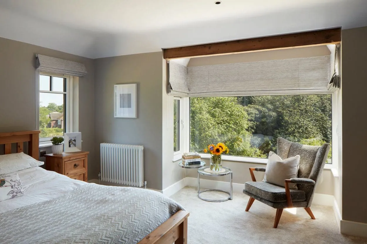 Textured oatmeal Roman blinds on a bay window and side window in a bedroom with an armchair