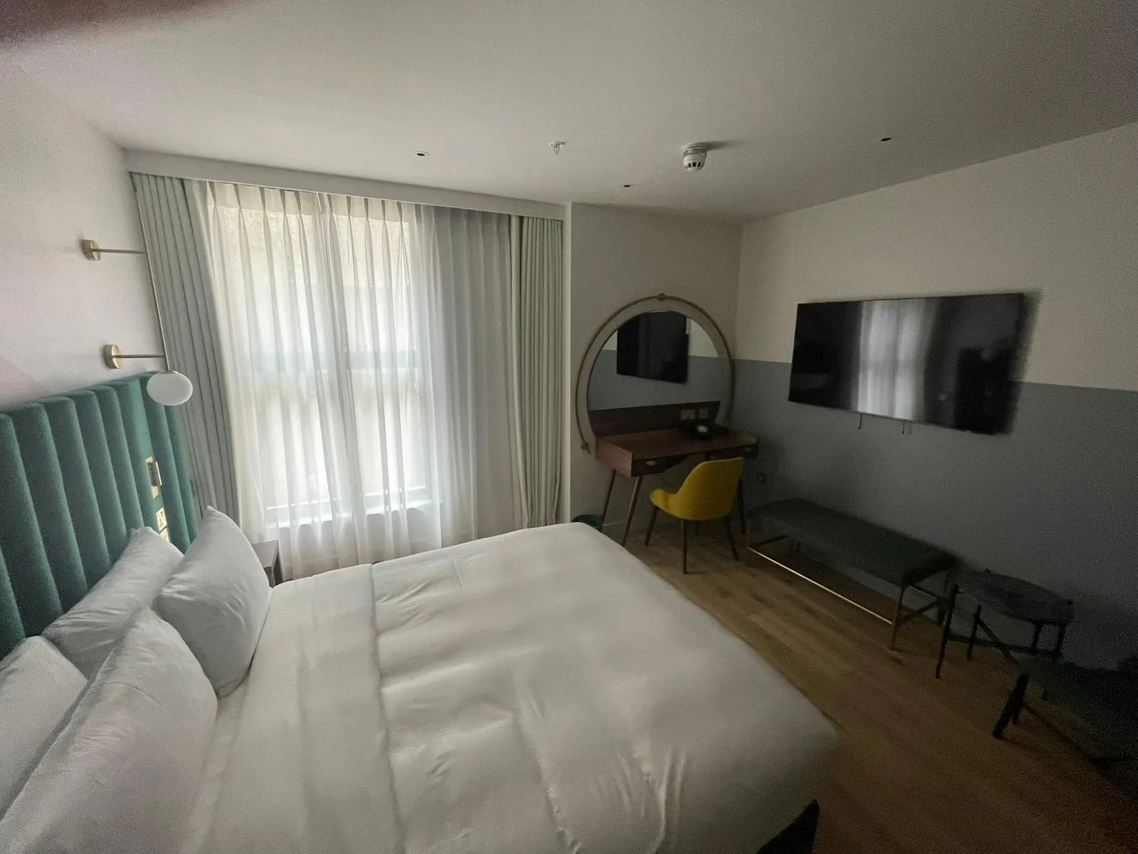 Sheer curtains at a hotel bedroom window, with a desk, round mirror and wall-mounted TV